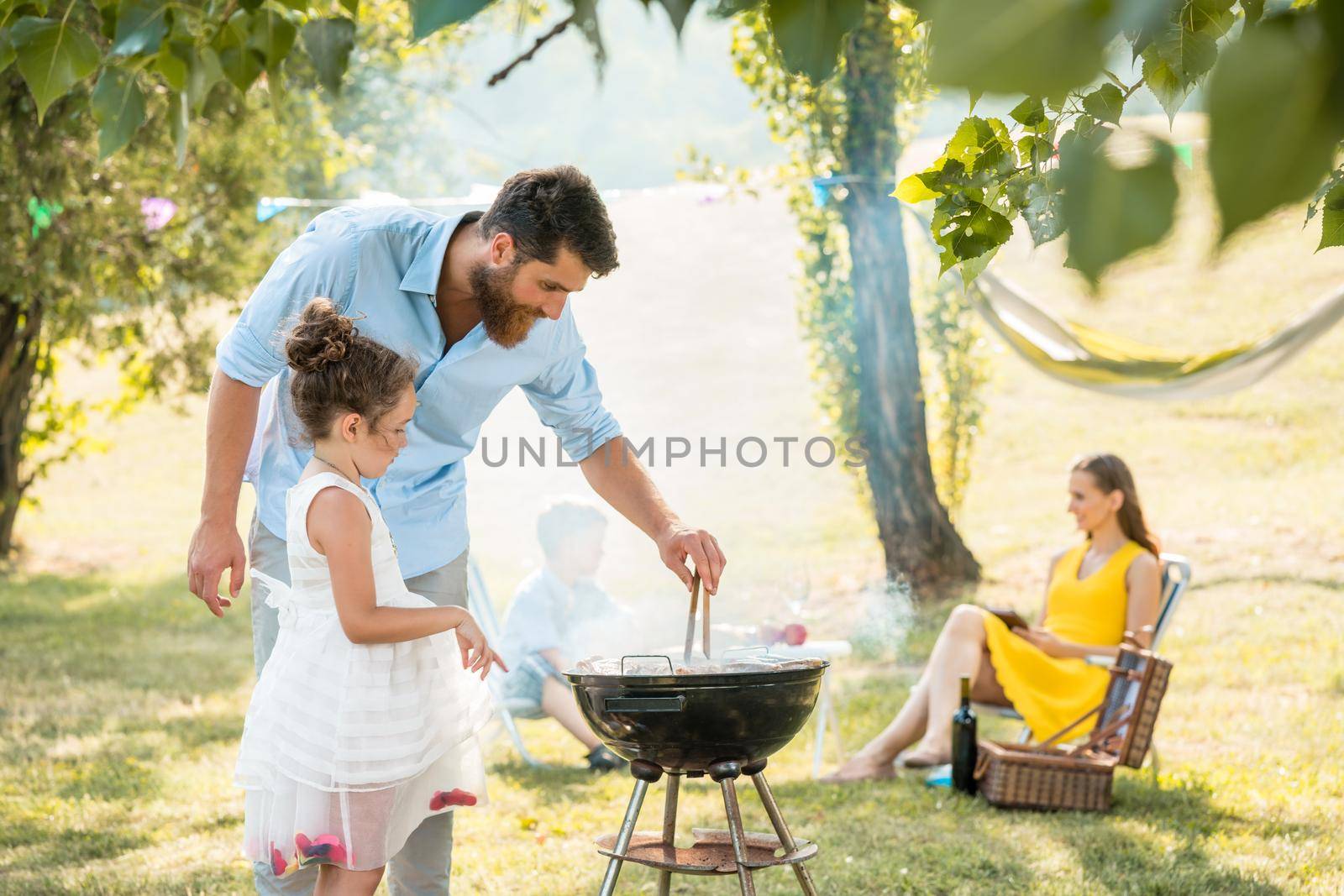 Girl watching father preparing meat on barbecue grill during family picnic by Kzenon