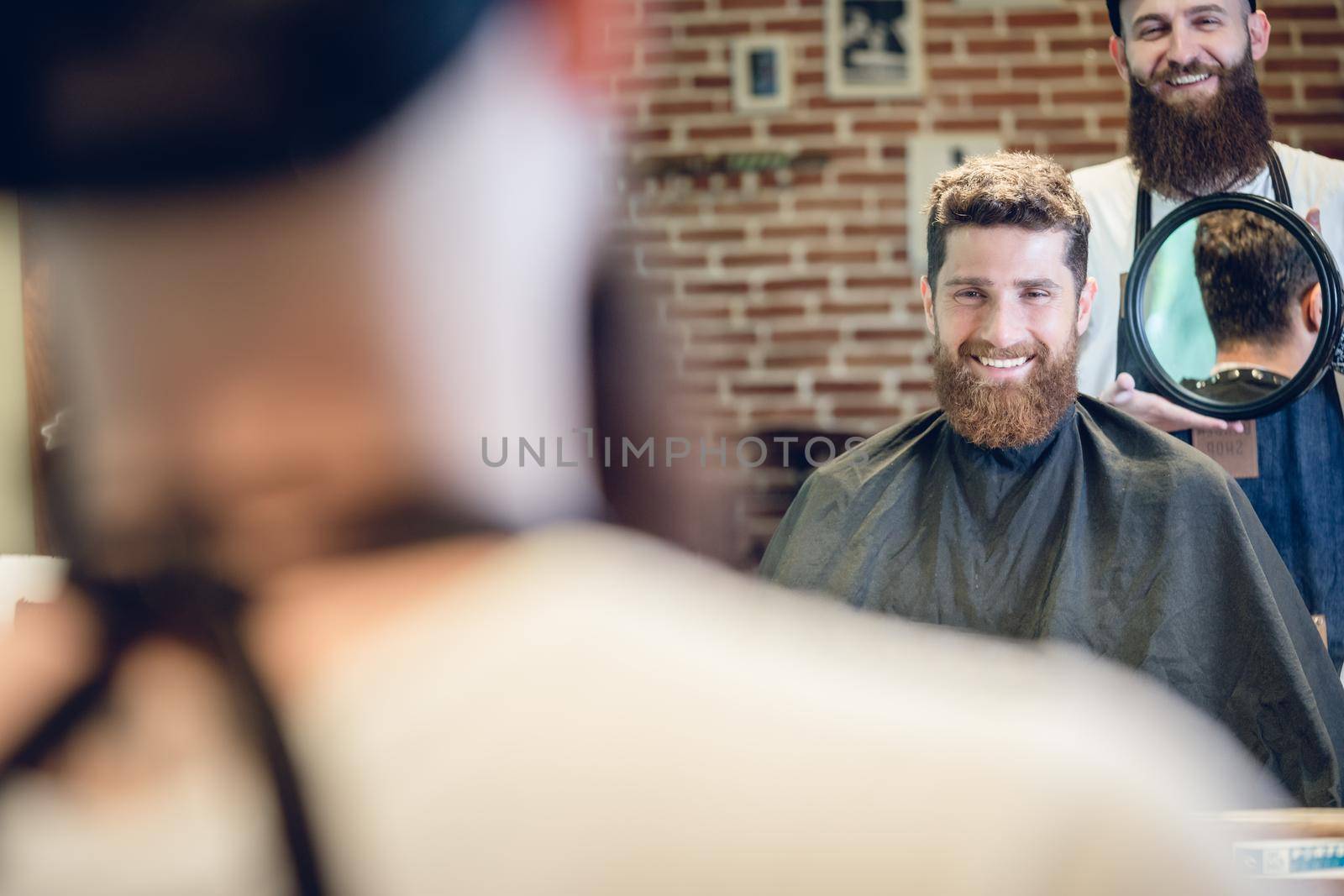 Over the shoulder view of a handsome young man smiling while looking at his new trendy haircut, in the mirror held by his experienced barber in a cool hair salon