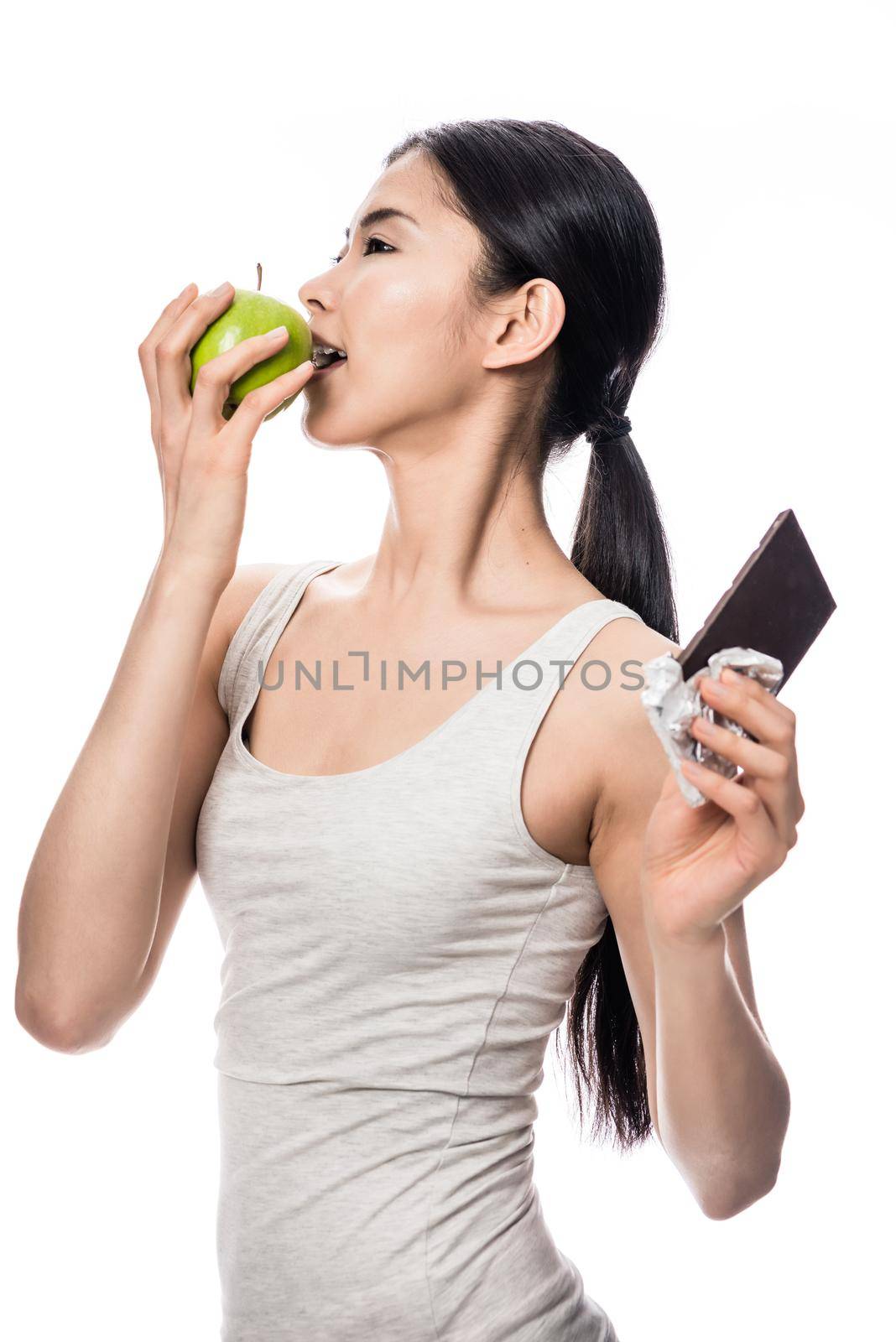 Attractive Asian woman opting for a healthy diet turning to bite into a fresh green apple while holding an unwrapped bar of chocolate aside
