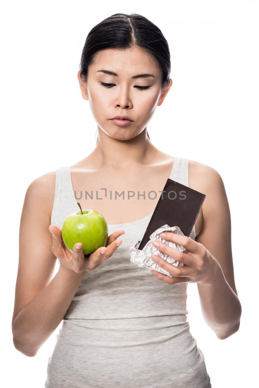 Pretty Asian woman debating an apple or chocolate as she stands with a fresh green apple in one hand and unwrapped bar of candy in the other in a healthy diet concept