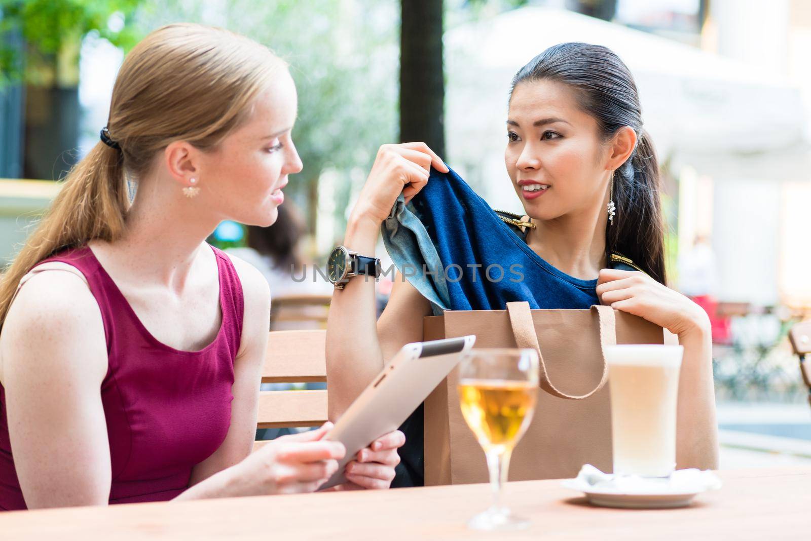 Two young women discussing a clothing purchase as they sit at a restaurant table ordering a meal as an attractive Asian women pulls it from the bag.