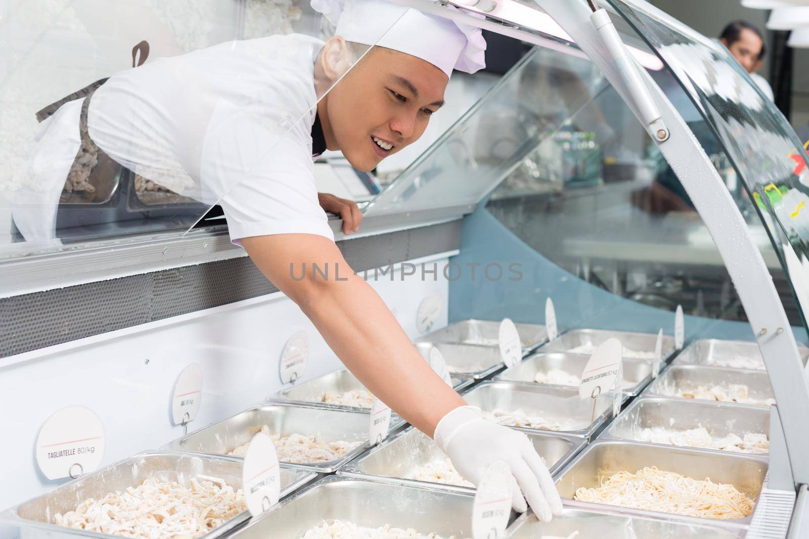 Asian chef placing food in trays into a glass counter in a delicatessen or restaurant buffet reaching forwards with a gloved hand to position it