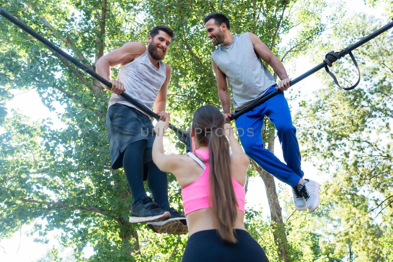 Low-angle rear view of a fit woman showing thumbs up to her two male friends during extreme workout outdoors in a modern calisthenics park