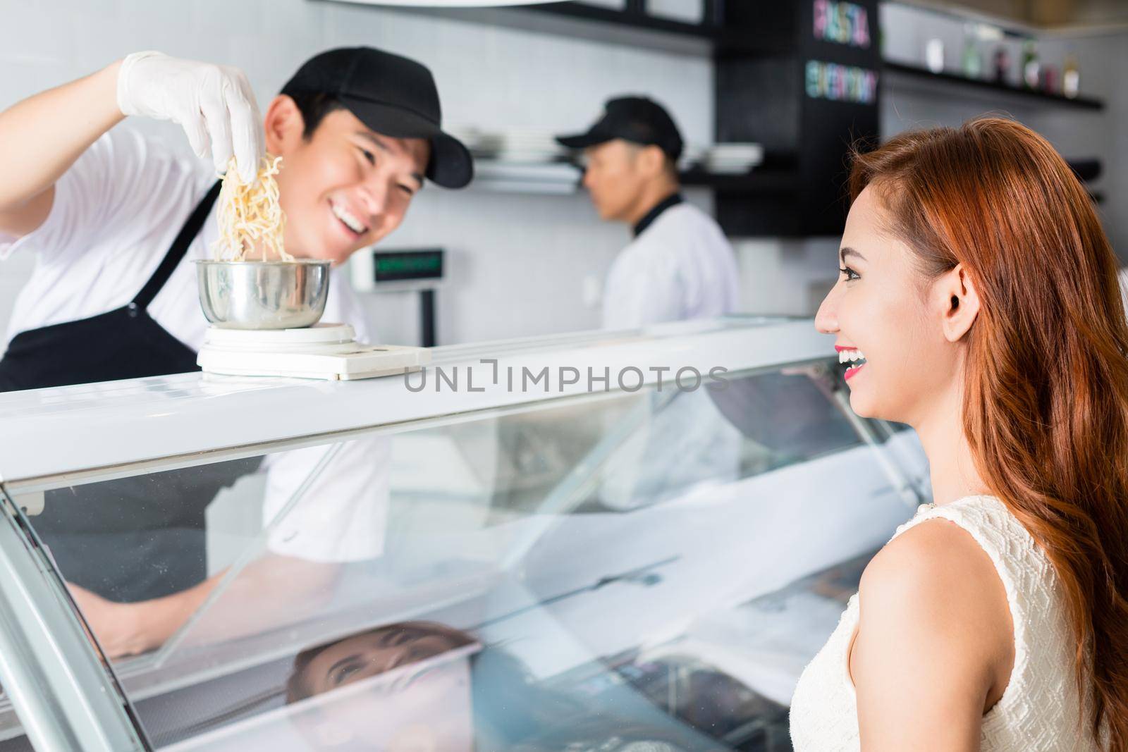 Laughing young Asian man serving a pretty woman weighing cheese at a delicatessen counter with a friendly smile