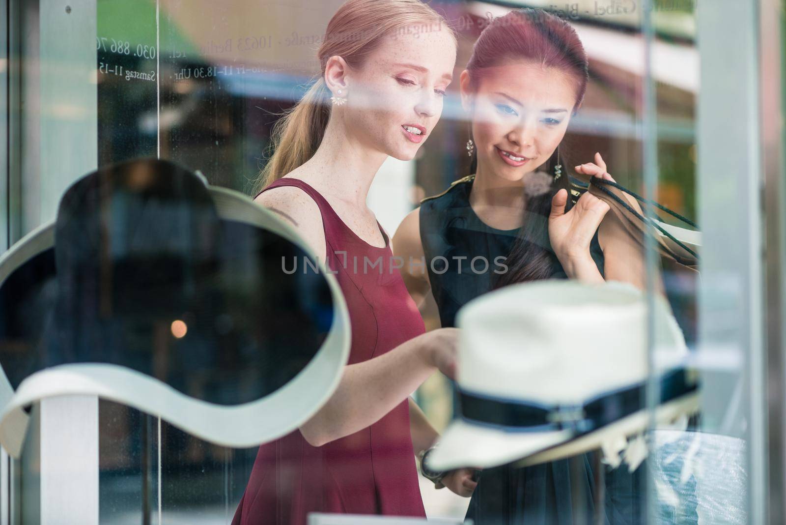 Two pretty young women window shopping pointing through the glass at a display of hats inside the fashion store , reflections on the glass