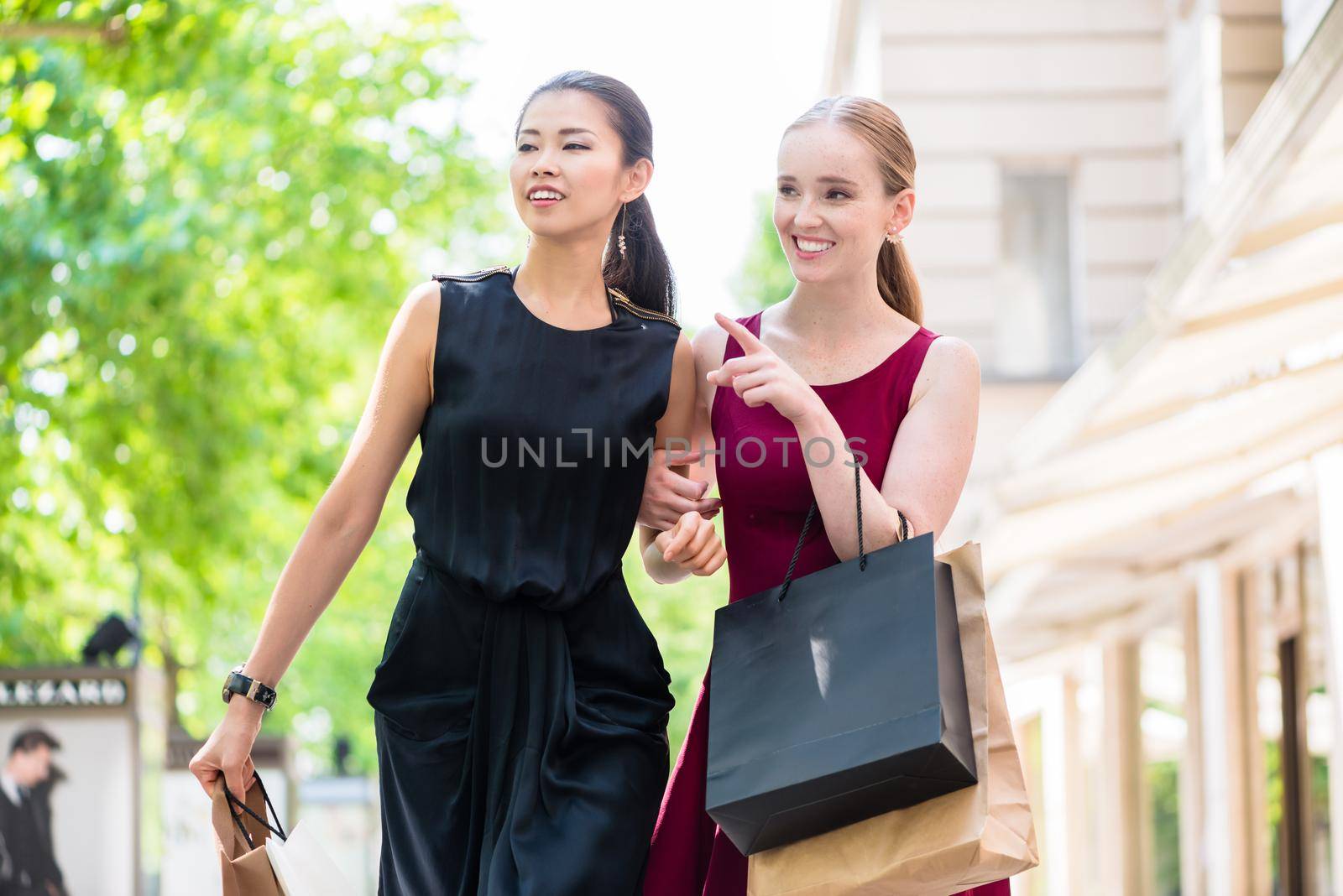 Two happy multiracial women out shopping together standing in an urban street pointing and smiling with bags over their arms