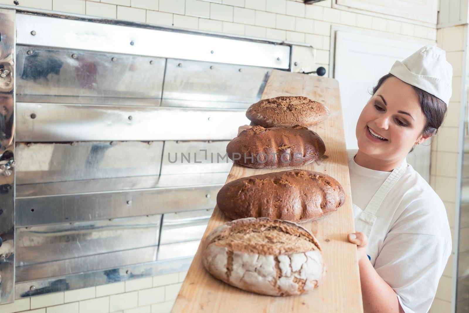 Baker woman presenting bread on board in bakery looking proudly into the camera