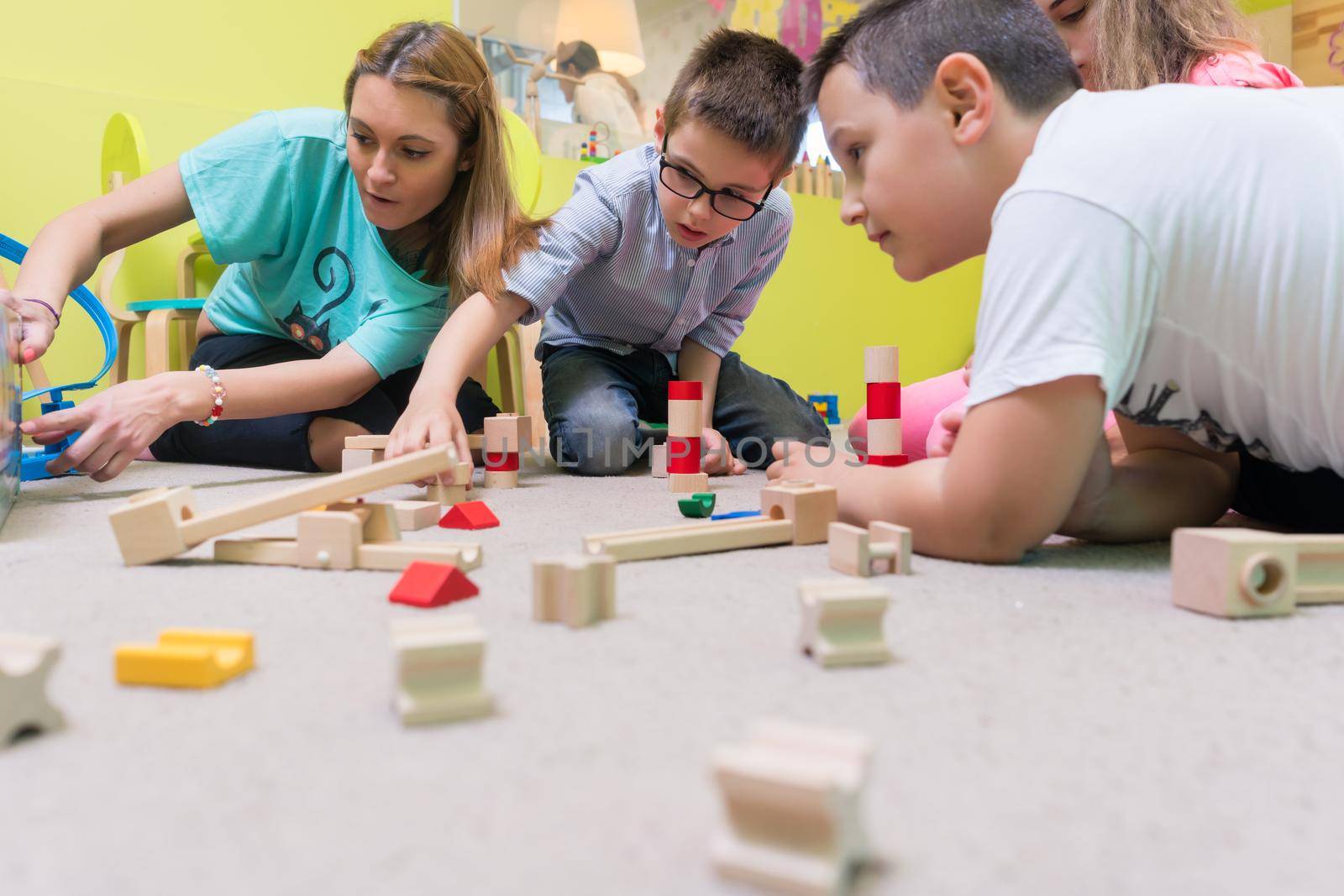 High-angle view of a young female educator teaching children to build with patience a wooden train circuit, during playtime on the floor in the kindergarten classroom