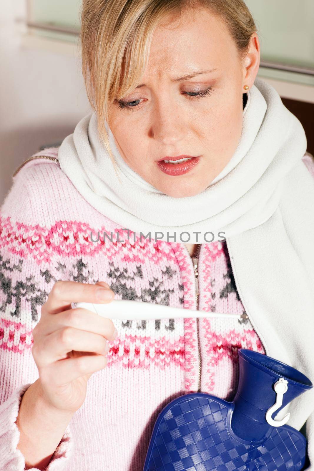 Woman with flu taking her temperature, looking at the thermometer having a hot water bag in her hand