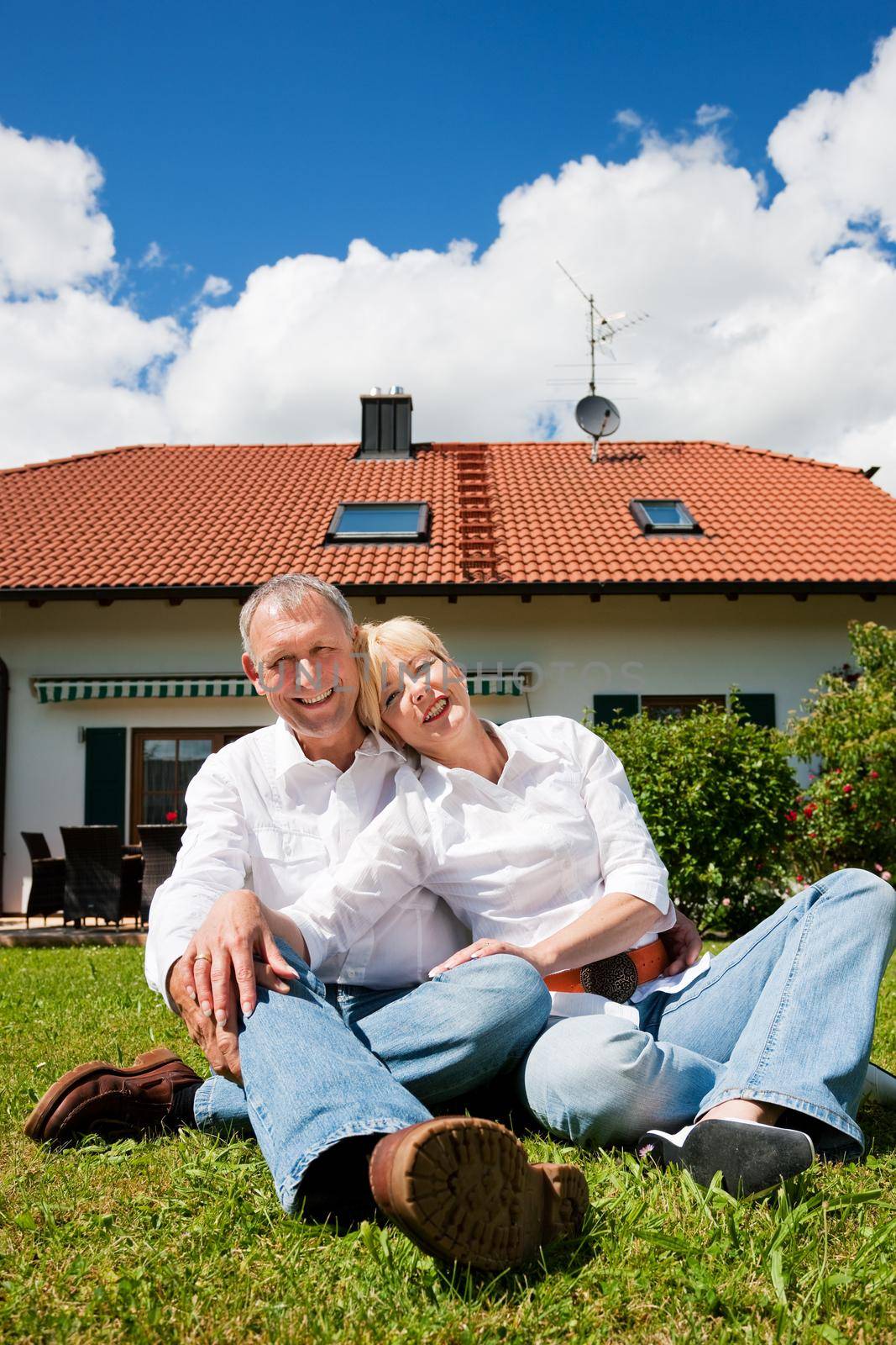 Senior couple sitting in the sun on the lawn in front of their new home - a detached house