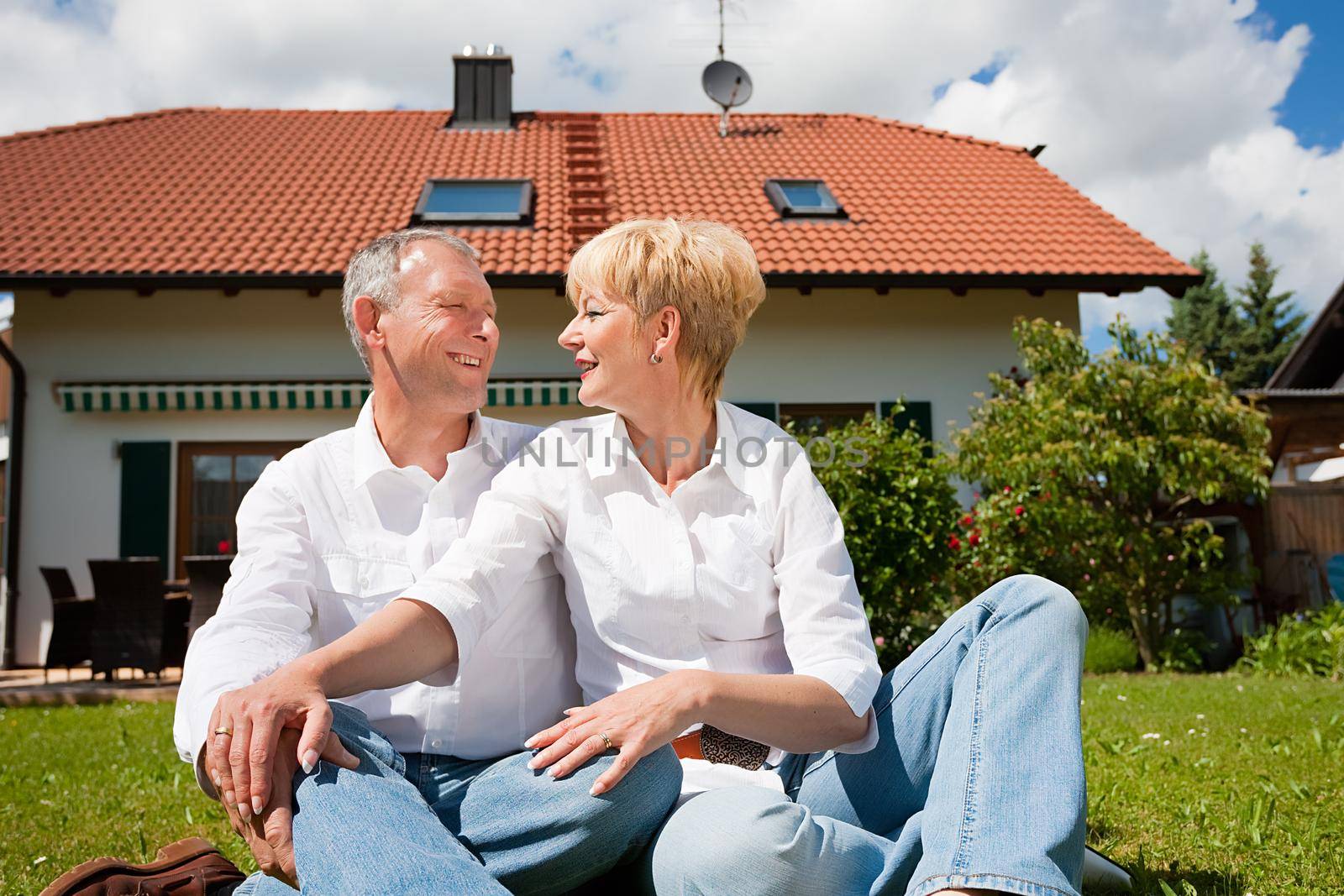 Senior couple sitting in the sun on the lawn in front of their new home - a detached house