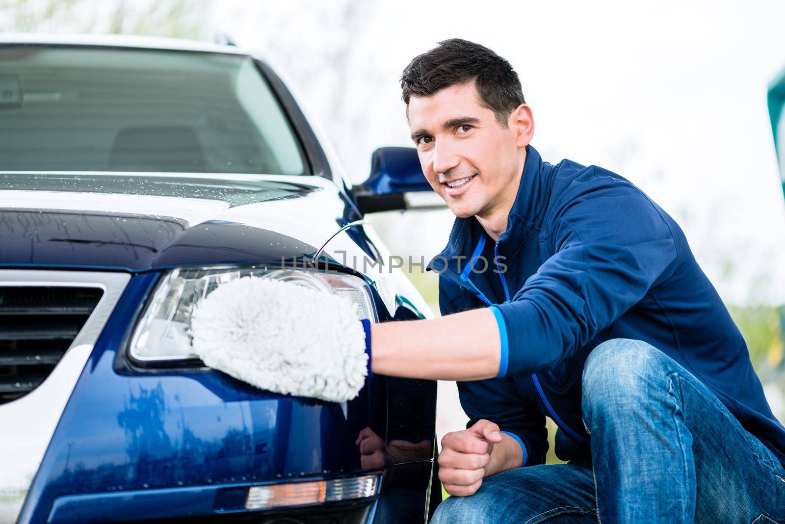 Smiling man cleaning the headlamp on his car wiping it with a mitt as he crouches alongside the vehicle