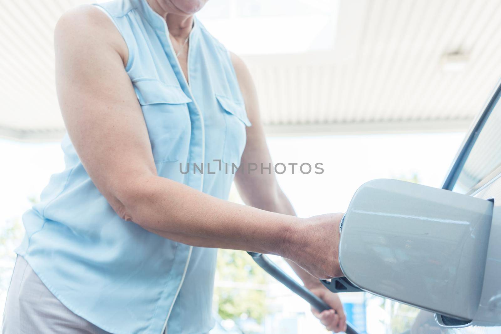Low-angle view portrait of an active senior woman smiling while filling up the gas tank of her car at the station in summer