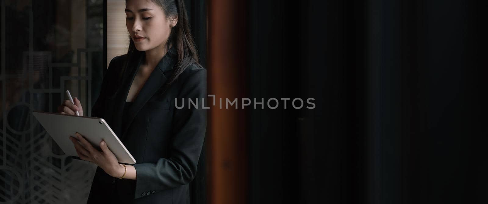 Attractive business asian woman using a digital tablet while standing in front of windows in an office building by nateemee