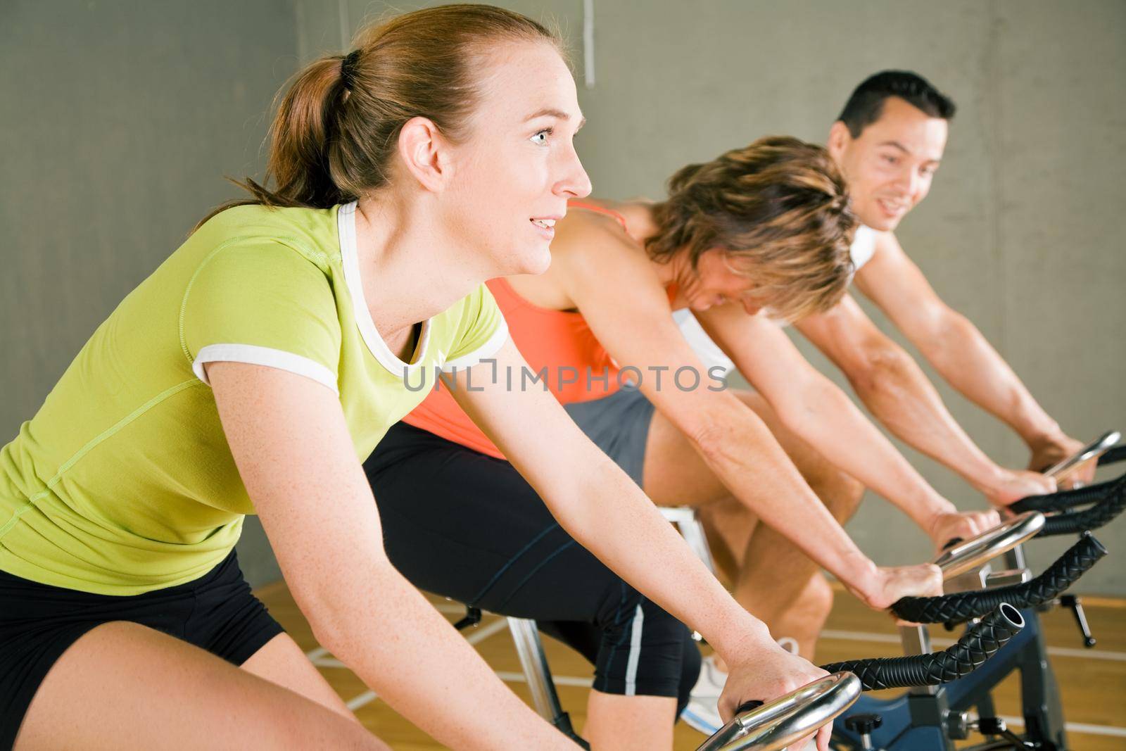 Three people cycling in a gym or fitness club, dressed in colorful clothes; focus on the girl in green in front