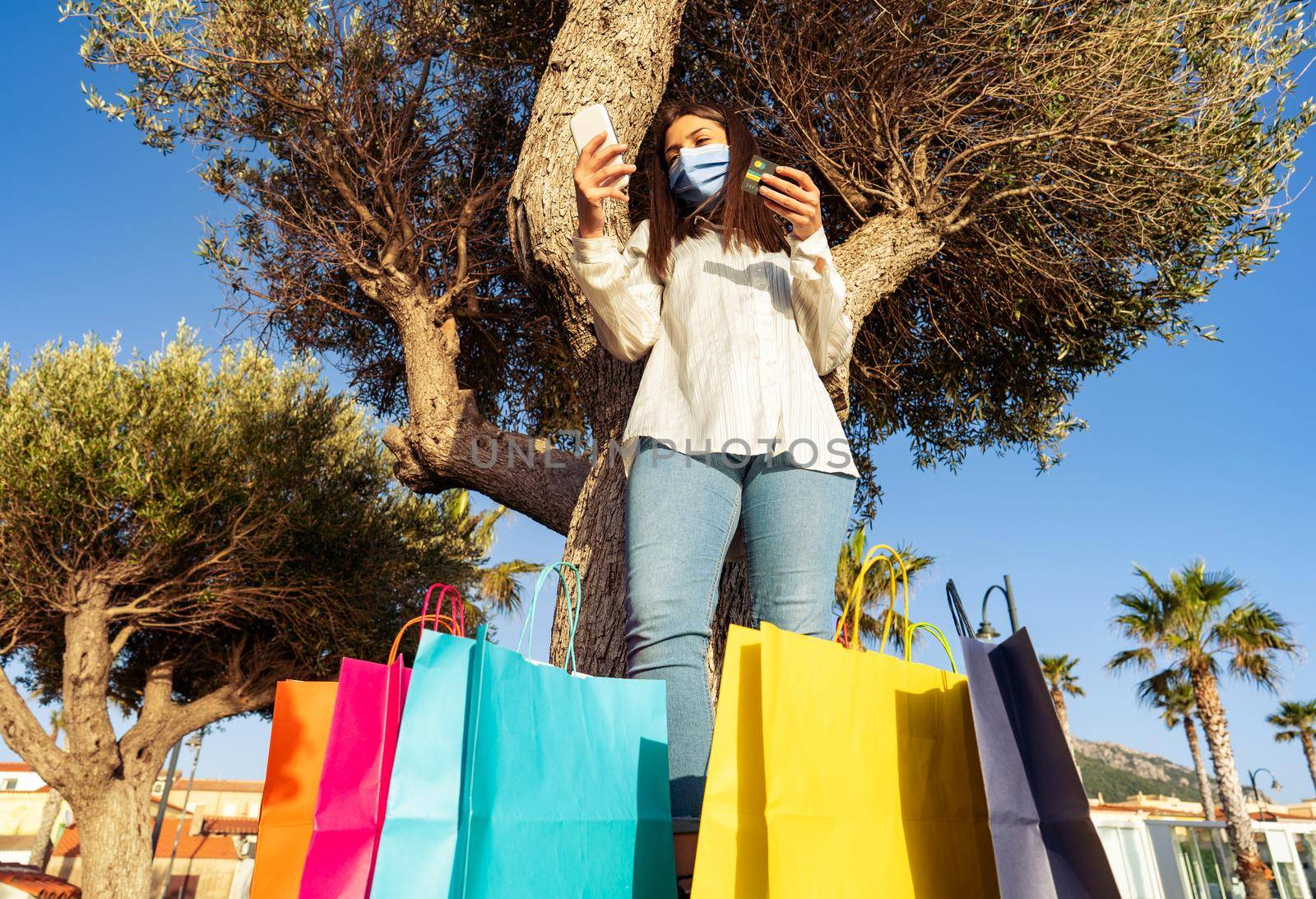 Lonely girl having fun in city park taking a break from a day of shopping using smartphone for new online credit card purchases. Young woman in Covid-19 protective face mask among many colored bags
