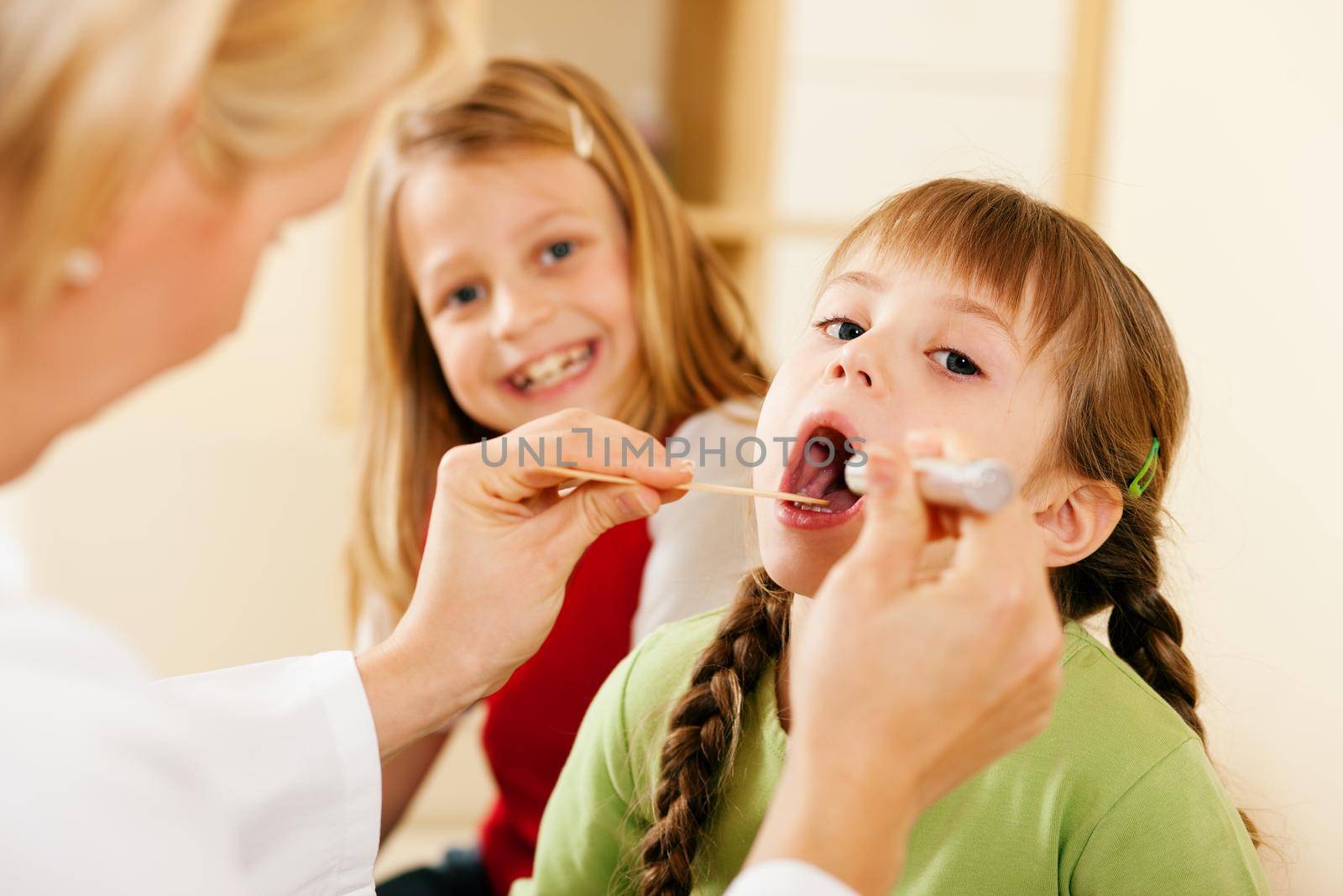 Female pediatrician doctor examining throat of a girl, in the background her sister is waiting to be examined as well