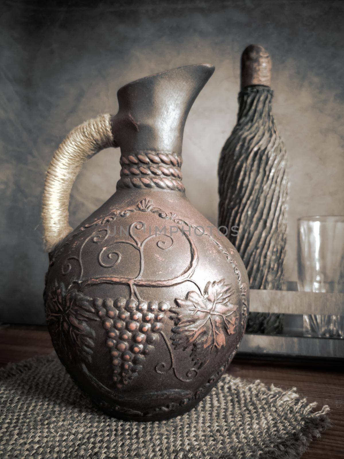 Ceramic wine jug made of red clay with a braided handle, handmade. There is a wine bottle and a glass next to it.