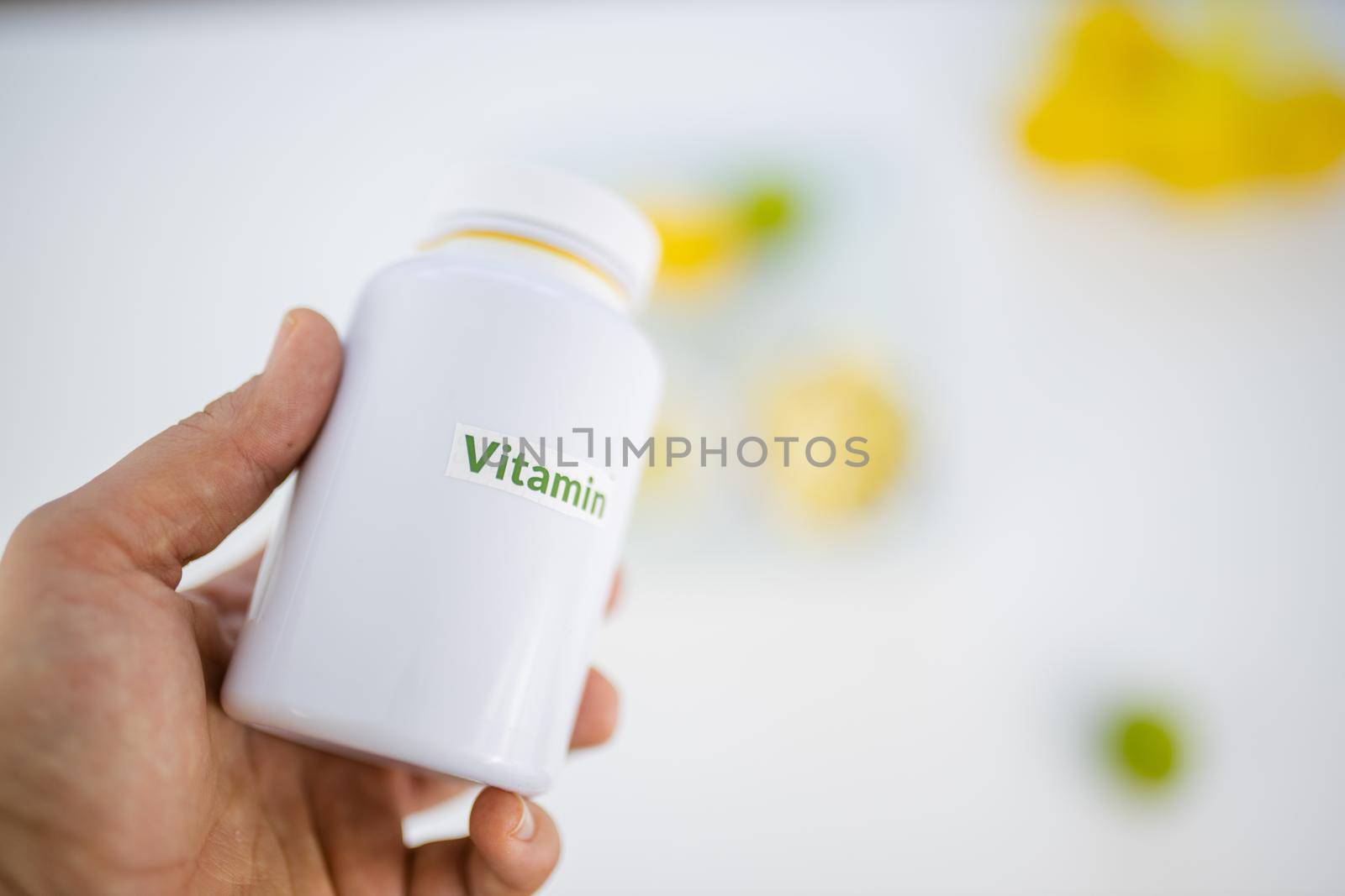 Hand holding plastic bottle of vitamins above blurry lemon and lime slices on white table. Hand holding white supplements bottle. Vitamin C nutrition
