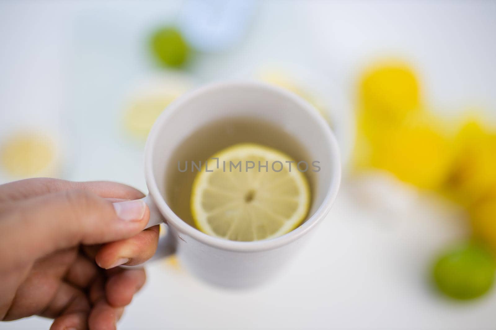 Female hand holding cup of lemon tea above blurry white table with limes and lemons. Woman holding white mug with lemon slice floating inside. Sweet citric beverages