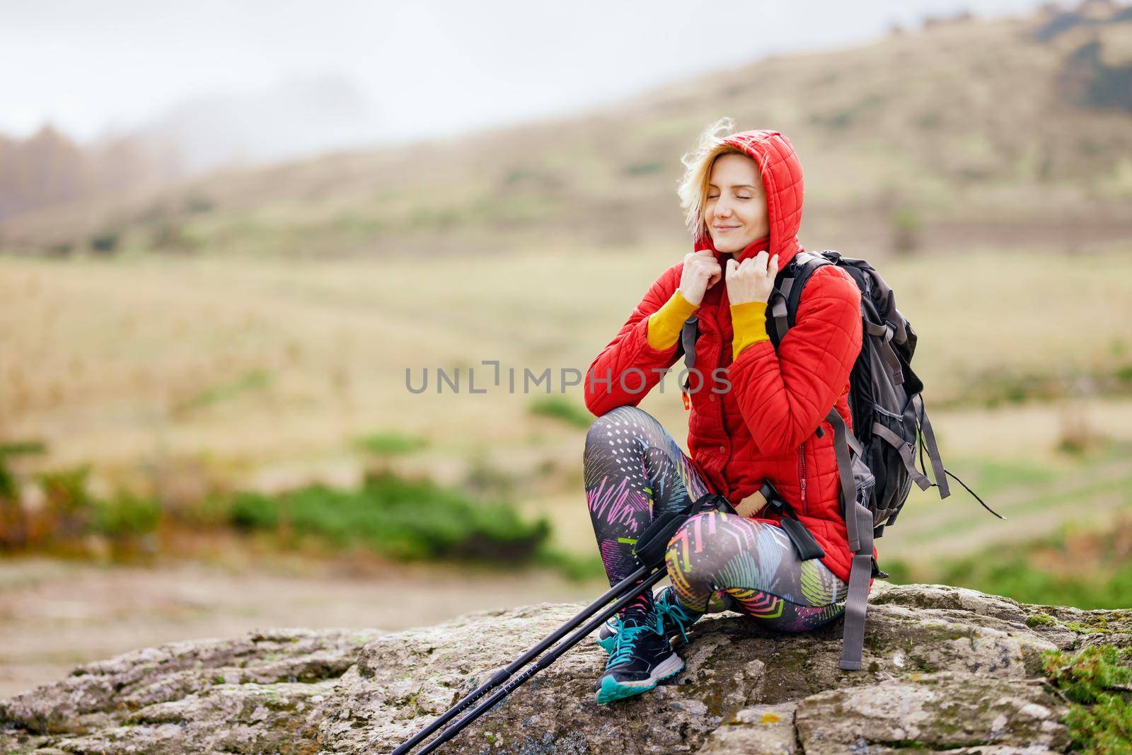 Hiker girl taking a rest on a rock in the mountains. Windy day. Travel and healthy lifestyle outdoors in fall season