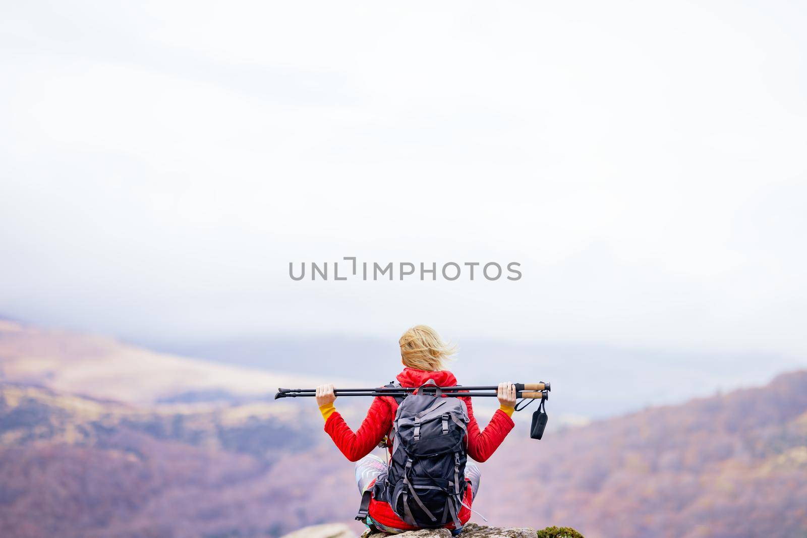 Hiker girl sitting on a rock in the mountains. Enjoying the view with hands up holding hiking poles. Travel and healthy lifestyle outdoors in fall season