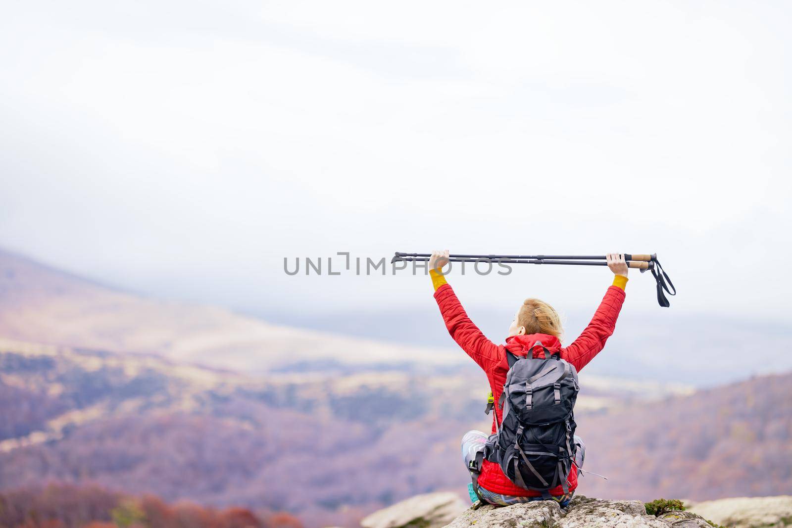 Hiker girl sitting on a rock in the mountains. Enjoying the view with hands up holding hiking poles. Travel and healthy lifestyle outdoors in fall season by kokimk