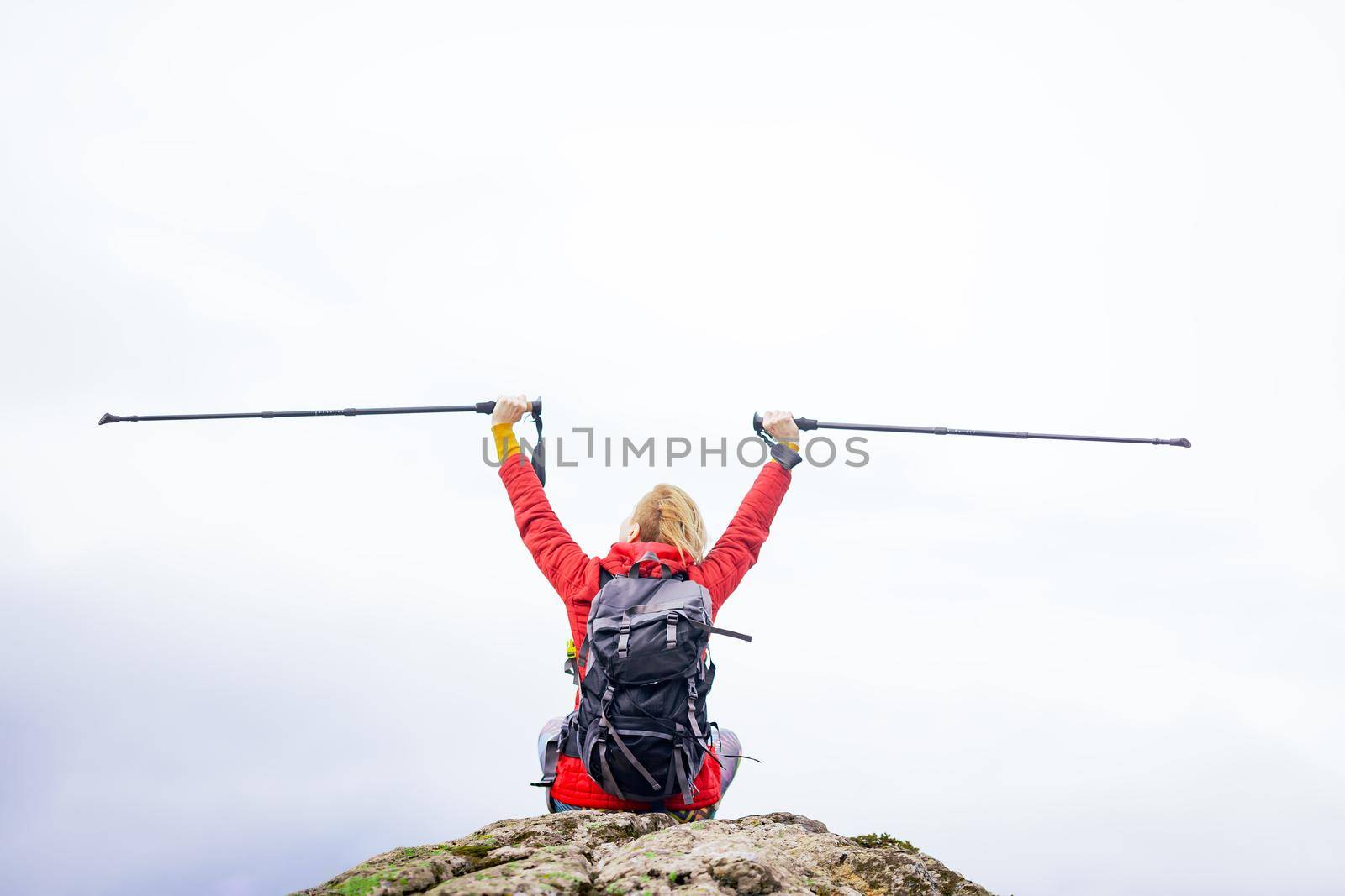 Hiker girl sitting on a rock in the mountains. Enjoying the view with hands up holding hiking poles. Travel and healthy lifestyle outdoors in fall season by kokimk