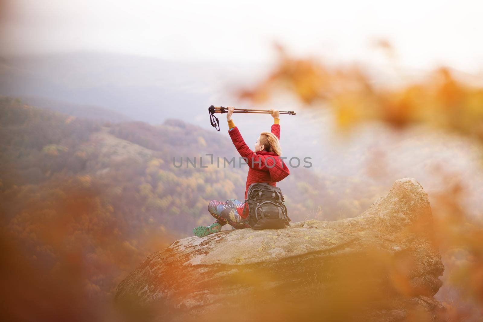 Hiker girl sitting on a rock in the mountains. Enjoying the view with hands up holding hiking poles. Travel and healthy lifestyle outdoors in fall season