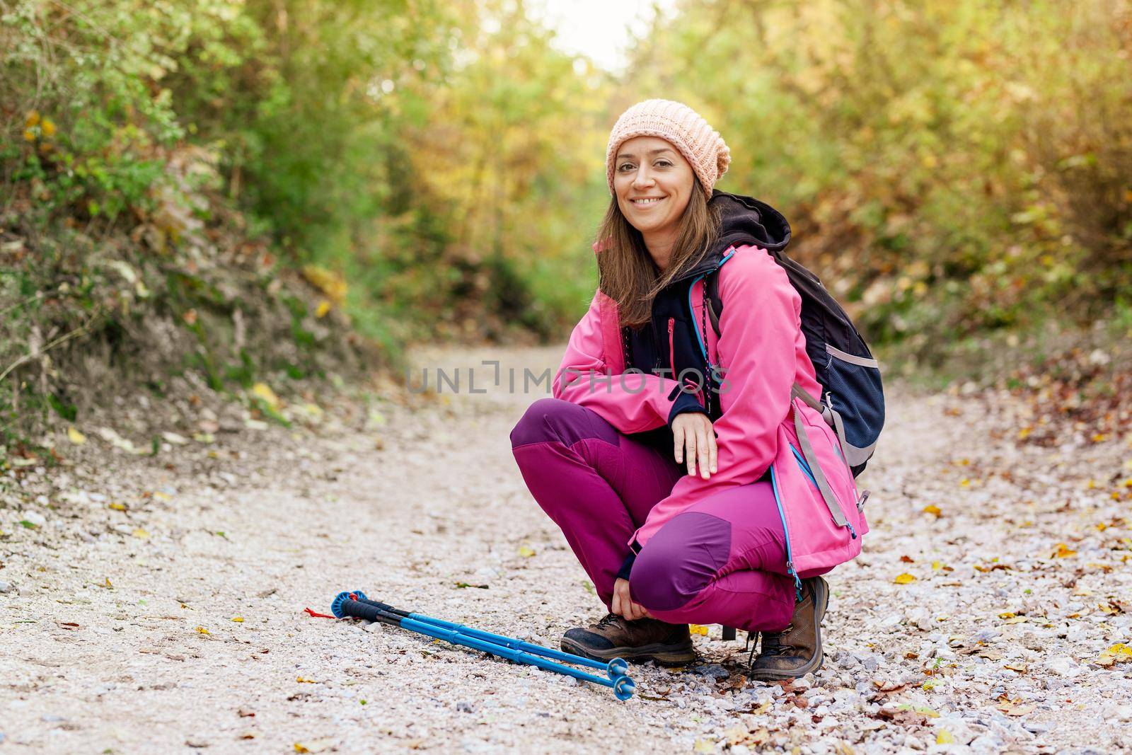 Hiker girl crouching on a wide trail in the mountains. Backpacker with pink jacket in a forest. Healthy fitness lifestyle outdoors.