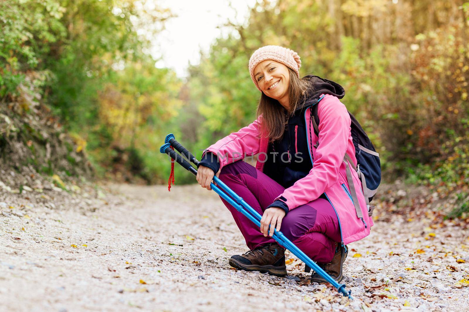 Hiker girl crouching on a wide trail in the mountains. Backpacker with pink jacket in a forest. Healthy fitness lifestyle outdoors.