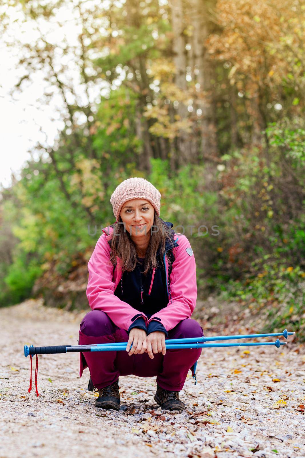 Hiker girl crouching on a wide trail in the mountains. Backpacker with pink jacket in a forest. Healthy fitness lifestyle outdoors. by kokimk