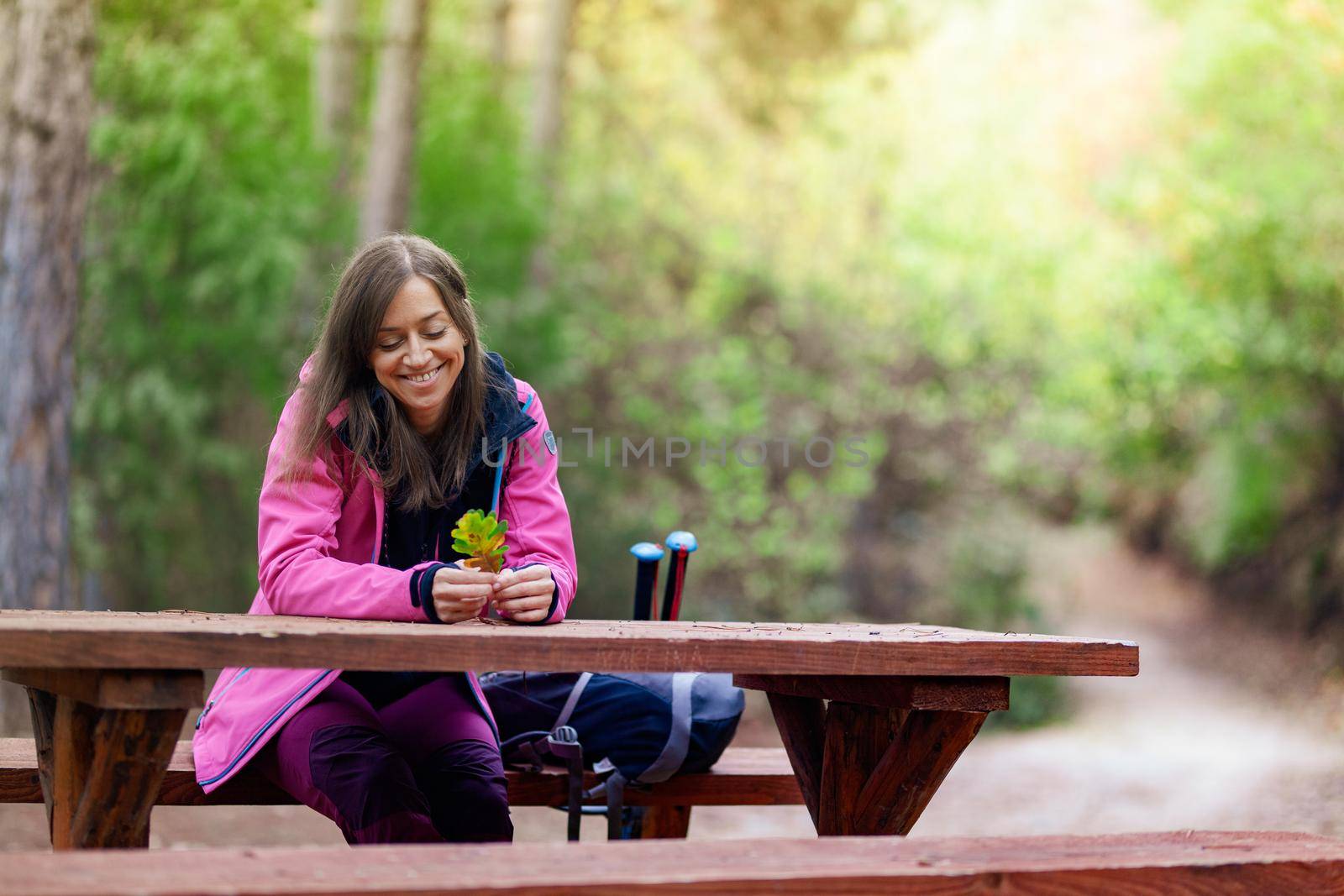 Hiker girl resting on a bench in the forest. Backpacker with pink jacket holding dry autumn leaves. by kokimk