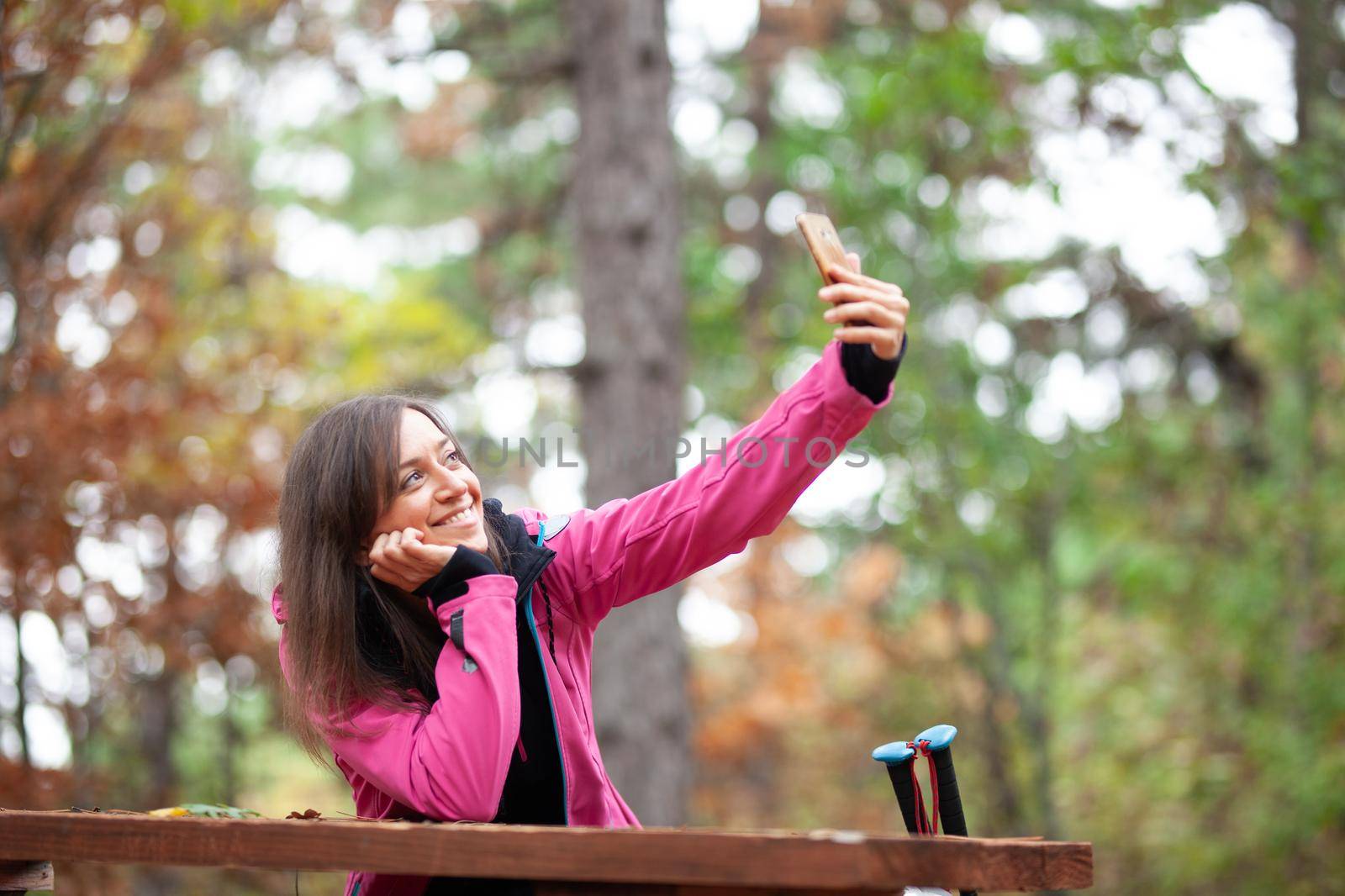Hiker girl resting on a bench in the forest. Backpacker with pink jacket taking selfie with smartphone. by kokimk