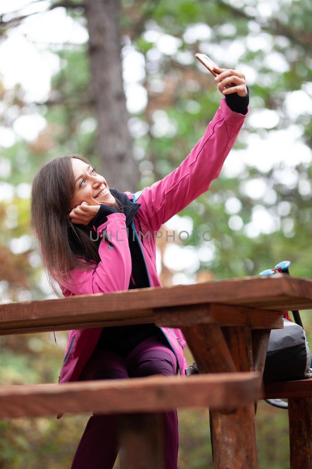 Hiker girl resting on a bench in the forest. Backpacker with pink jacket taking selfie with smartphone. by kokimk