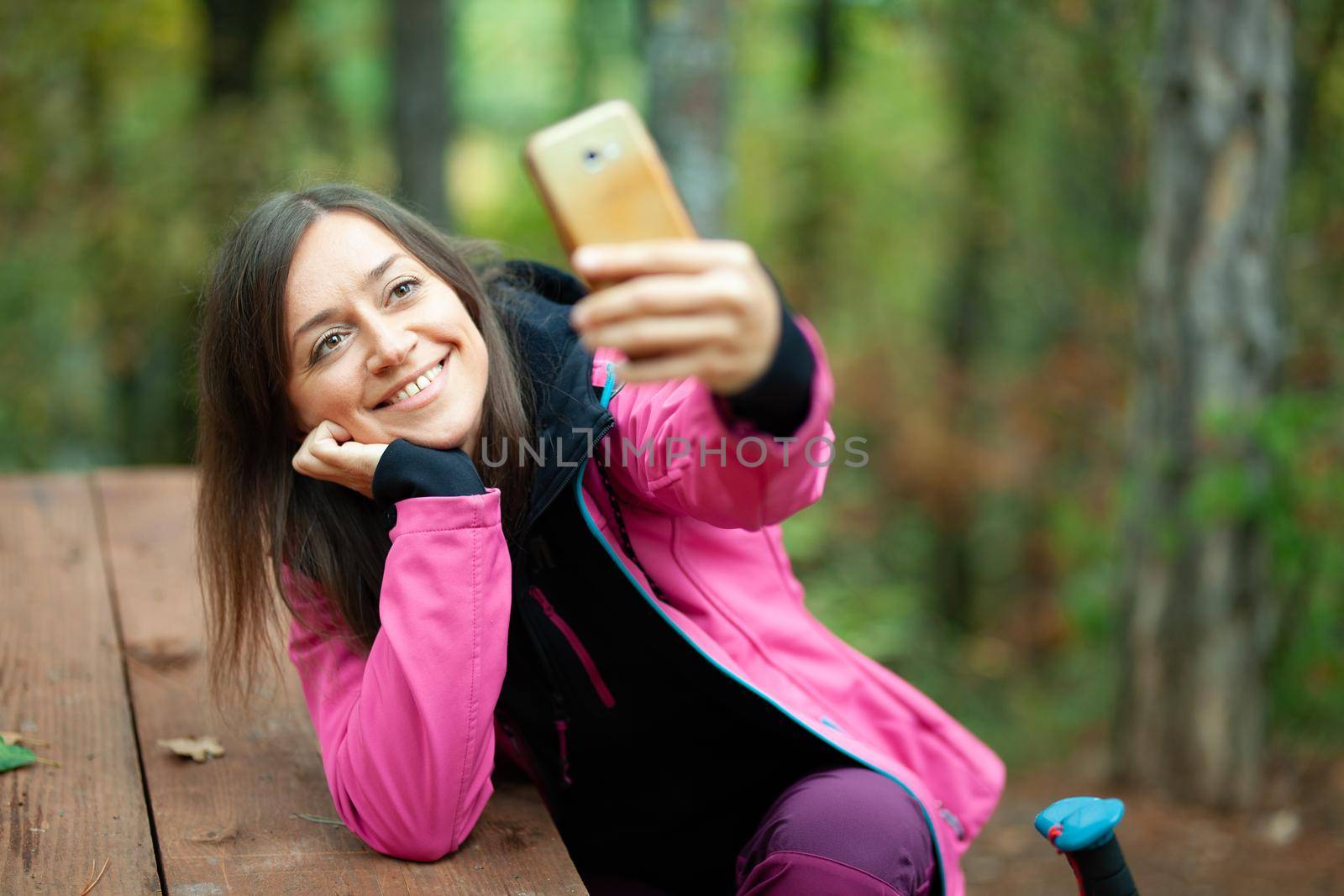 Hiker girl resting on a bench in the forest. Backpacker with pink jacket taking selfie with smartphone. by kokimk