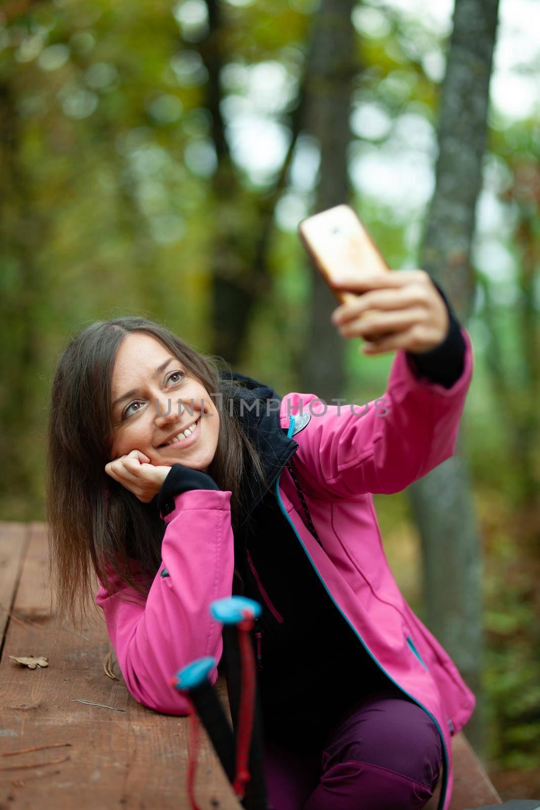 Hiker girl resting on a bench in the forest. Backpacker with pink jacket taking selfie with smartphone.