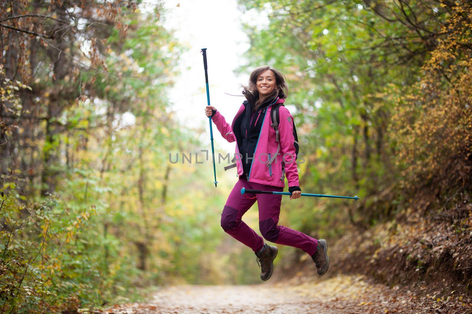 Hiker girl jumping on a trail in the mountains. Backpacker with hiking poles and pink jacket in a forest. Happy lifestyle outdoors.
