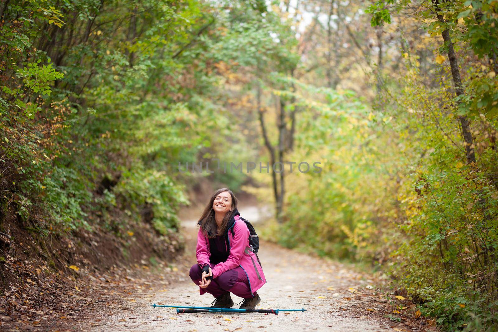 Hiker girl crouching on a trail in the mountains. Backpacker with pink jacket in a forest. Healthy fitness lifestyle outdoors. by kokimk