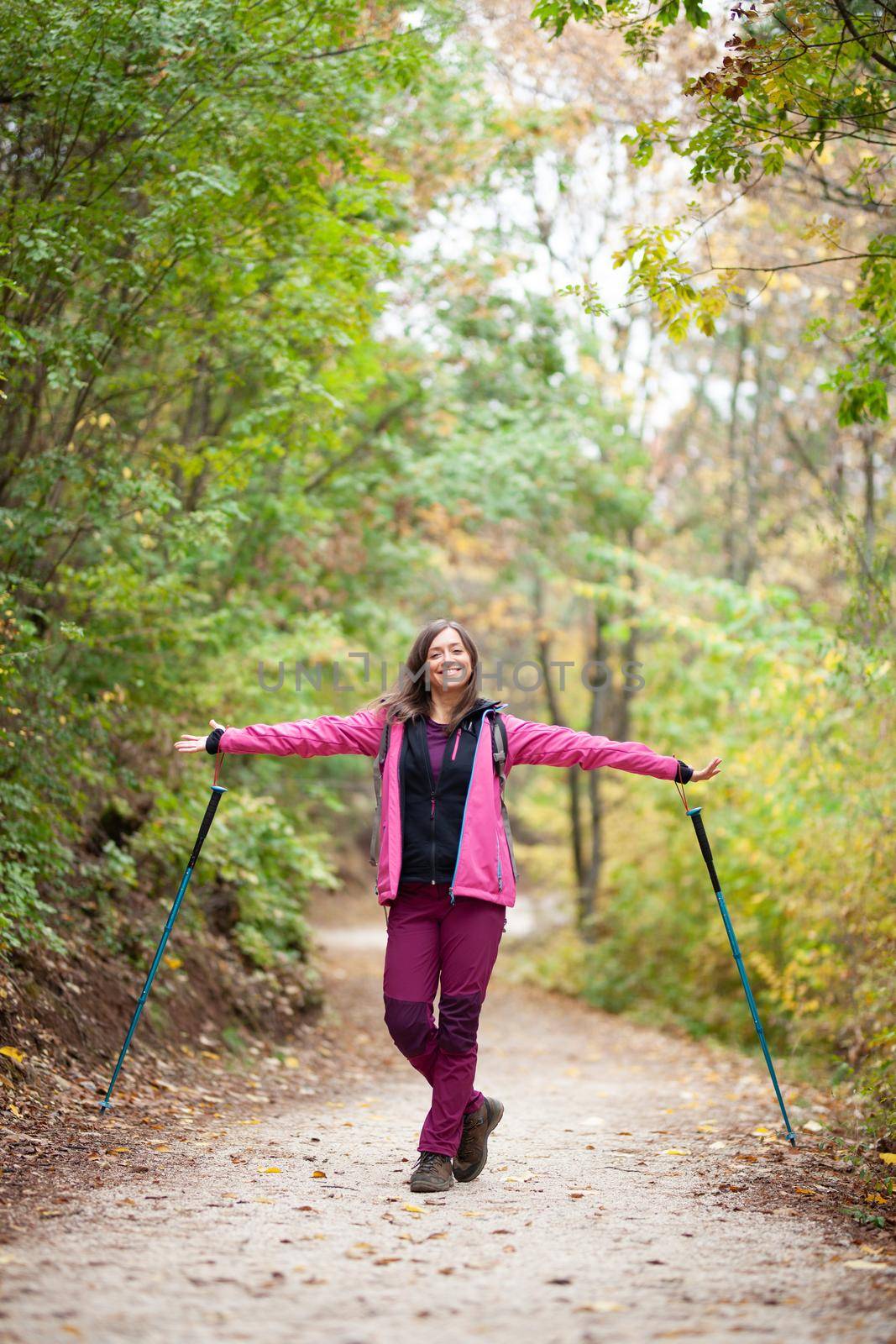 Hiker girl standing on a wide trail in the mountains. Backpacker with pink jacket in a forest. Healthy fitness lifestyle outdoors.