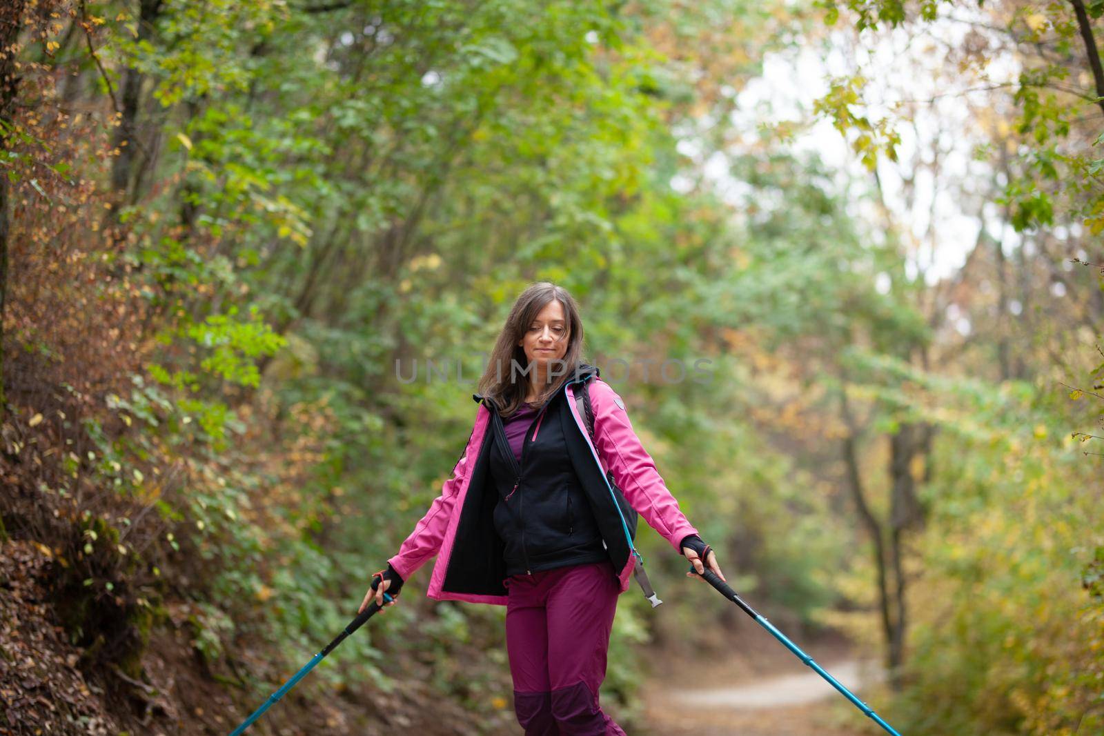 Hiker girl jumping on a trail in the mountains. Backpacker with hiking poles and pink jacket in a forest. Happy lifestyle outdoors.