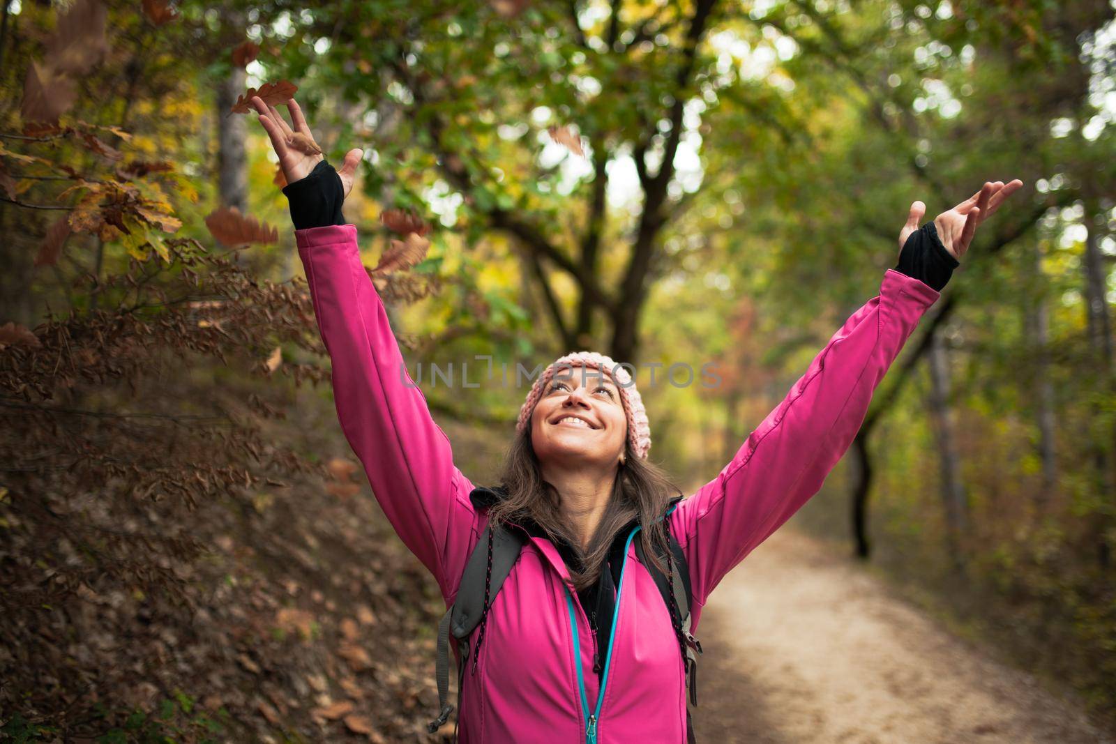 Hiking girl in pink on a trail in the forest. Hands up enjoying the falling leaves in nature in fall season.