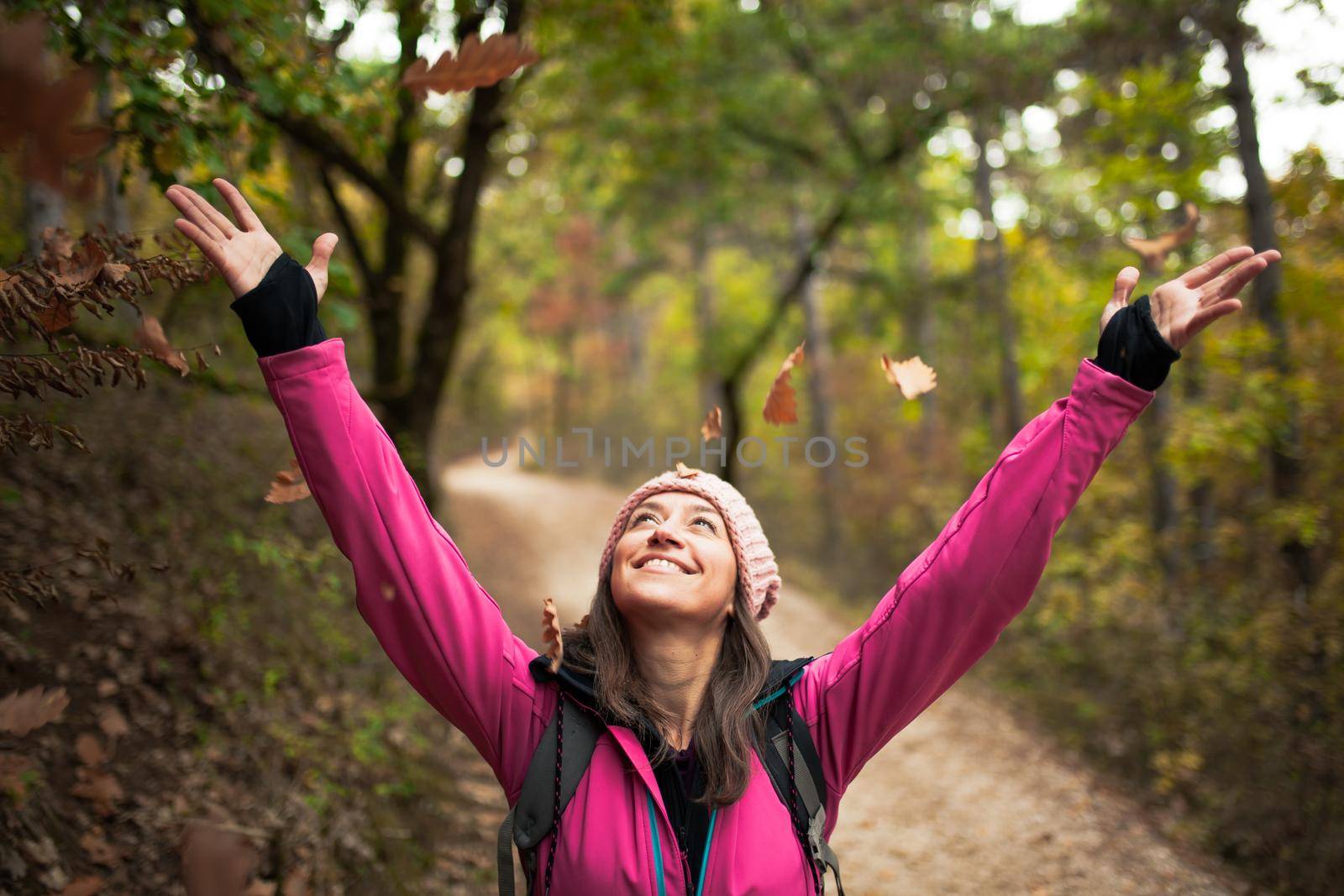 Hiking girl in pink on a trail in the forest. Hands up enjoying the falling leaves in nature in fall season.