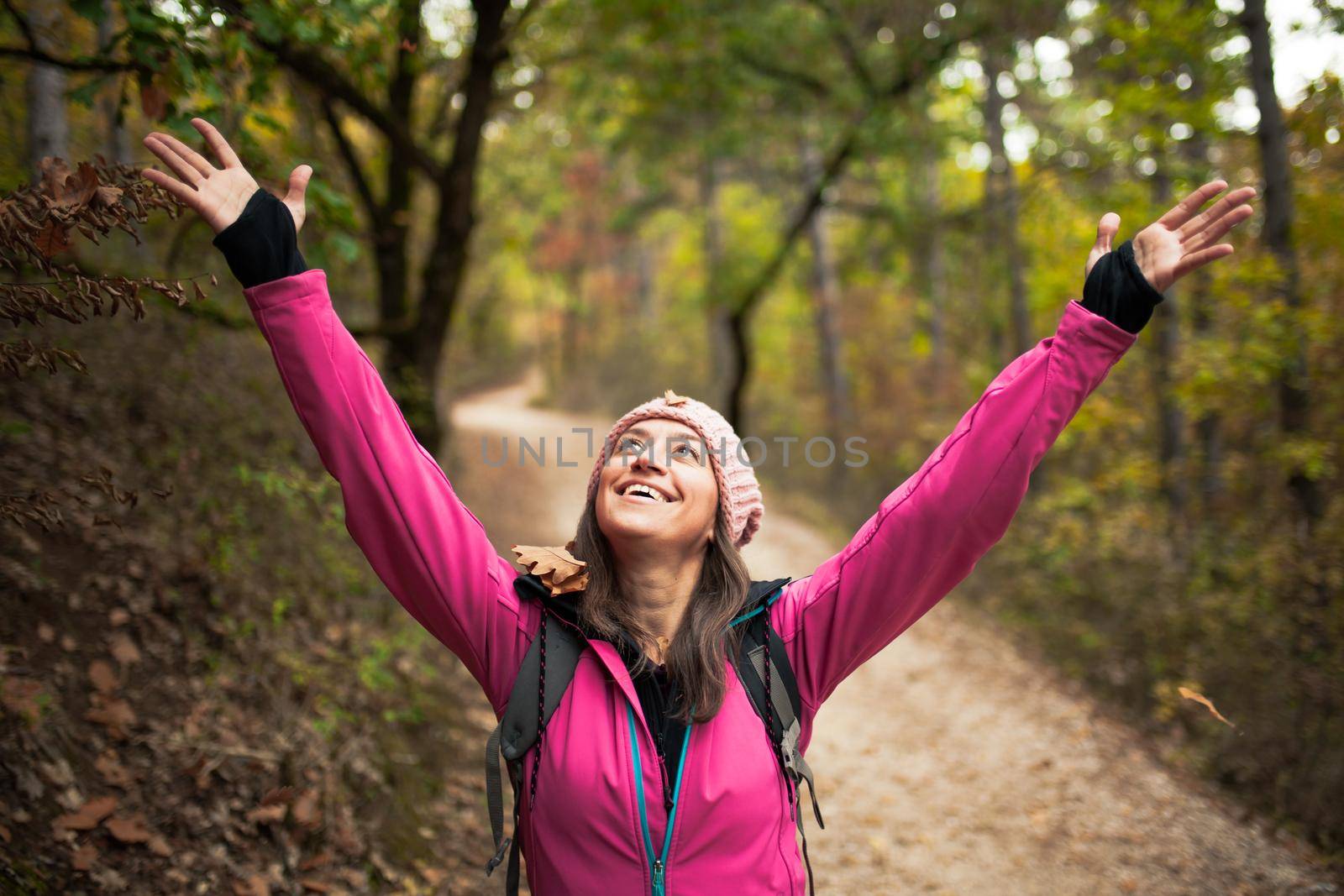 Hiking girl in pink on a trail in the forest. Hands up enjoying the falling leaves in nature in fall season. by kokimk