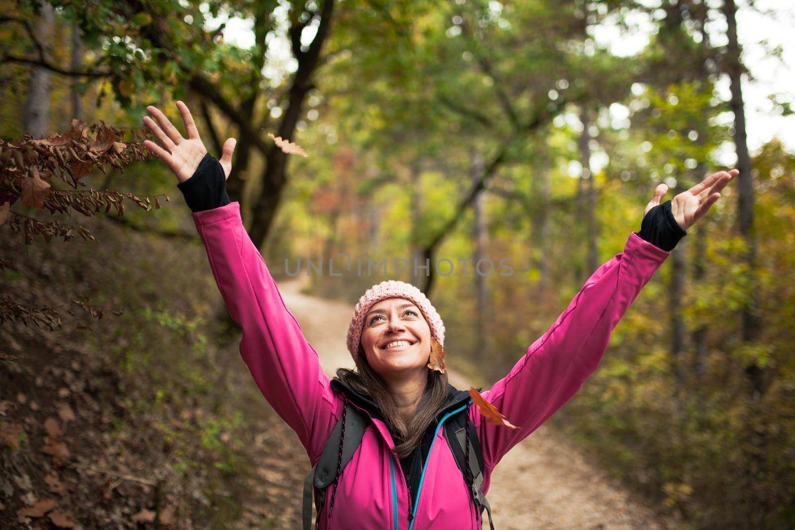 Hiking girl in pink on a trail in the forest. Hands up enjoying the falling leaves in nature in fall season.