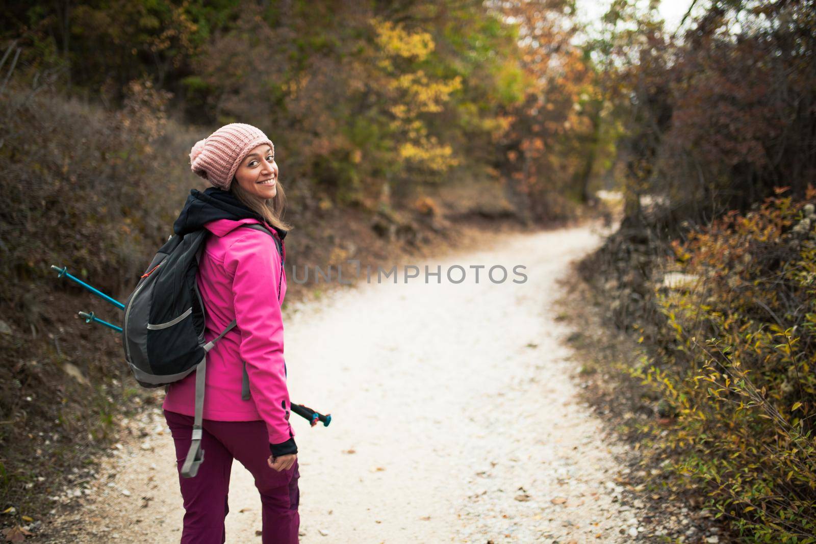 Hiker girl walking on a path in the mountains, looking back at the camera. Backpacker with pink jacket in a forest. Healthy fitness lifestyle outdoors.