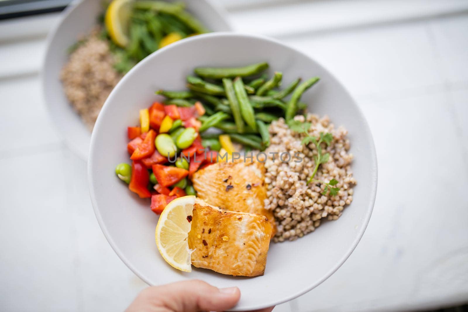 Hand holding salmon and buckwheat dish with green beans, broad beans, and tomato slices. Nutritious dish with vegetables and fish on white plate. Healthy balanced diet