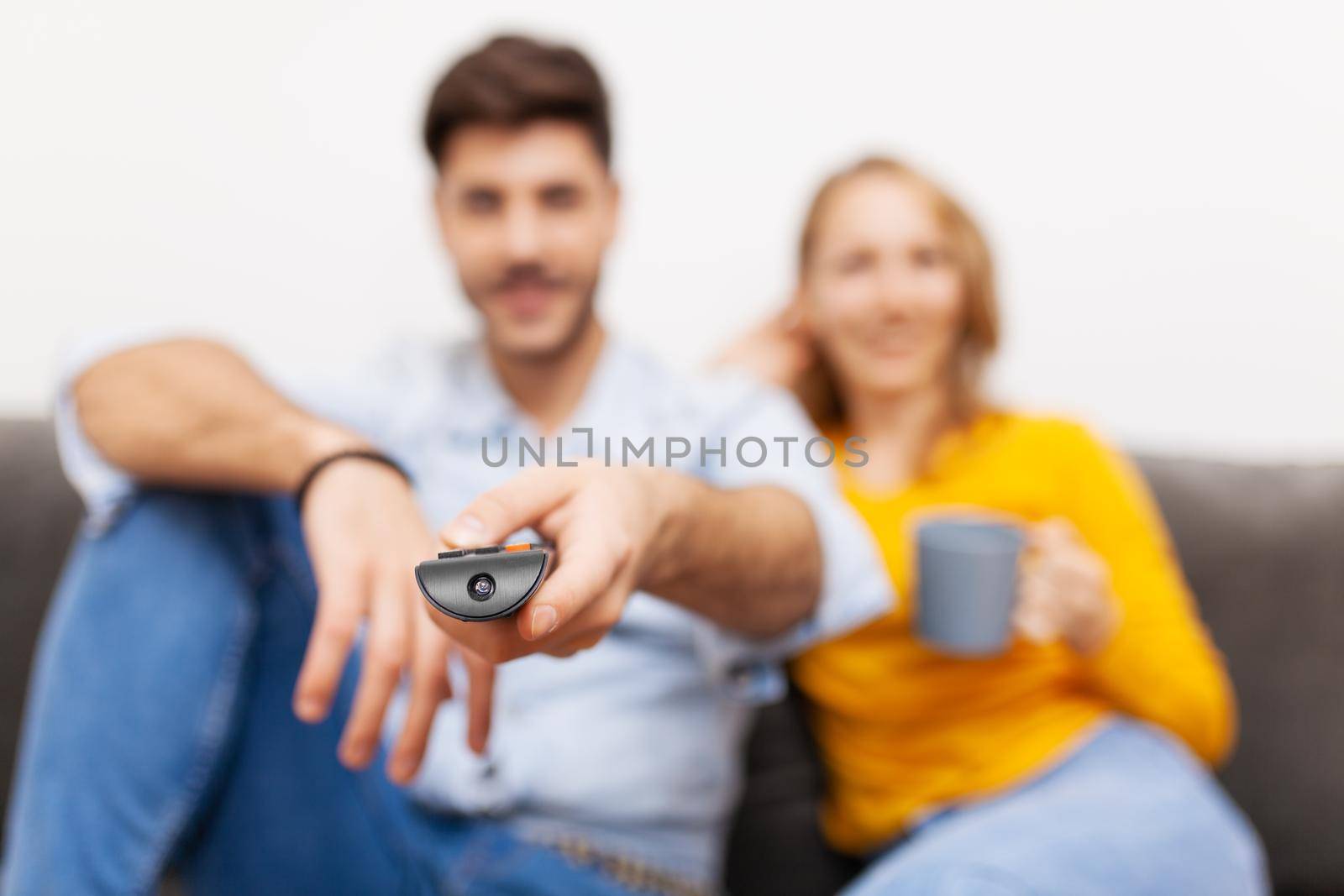 couple in love on a sofa watching tv, holding coffee and remote control by kokimk