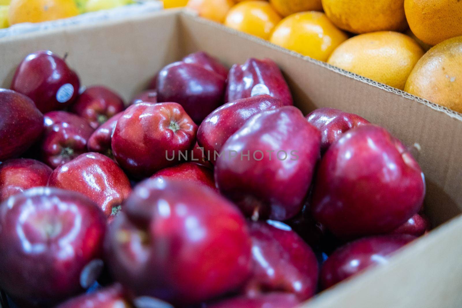 Close-up of fresh red apples in cardboard box next to pile of oranges. Juicy-looking fruit for sale on stand inside classic Mexican market. Natural healthy food