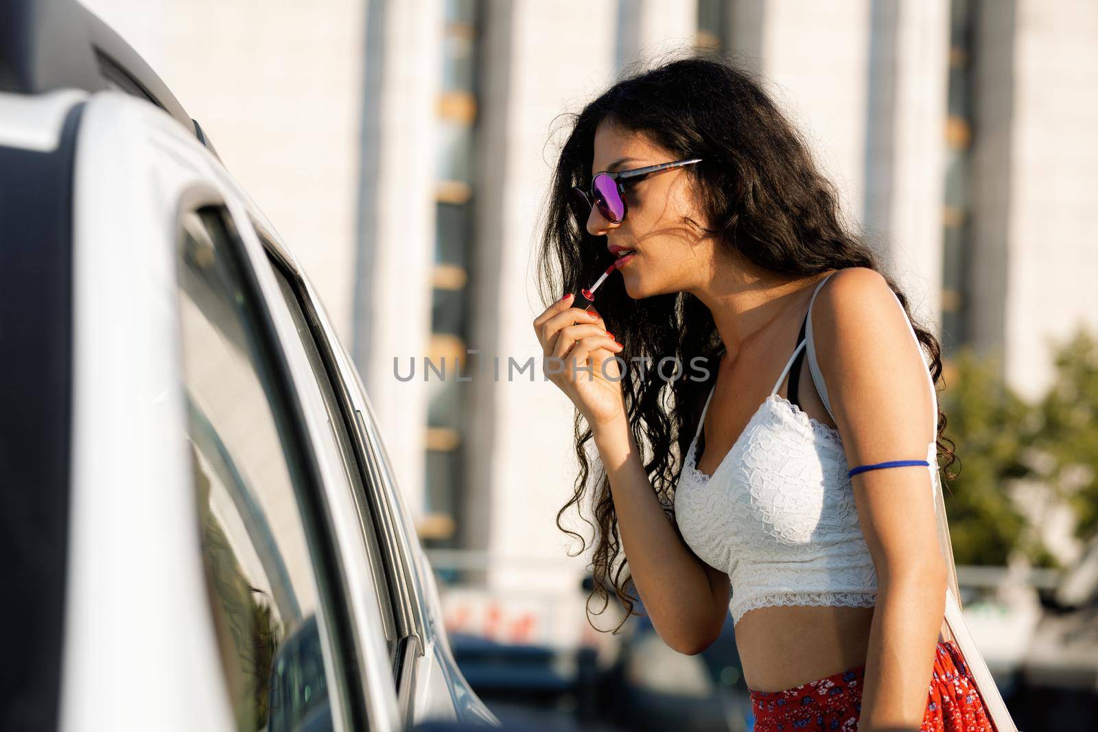 Beautiful girl with sunglasses putting on lipstick using a car mirror. Sunny summer day.
